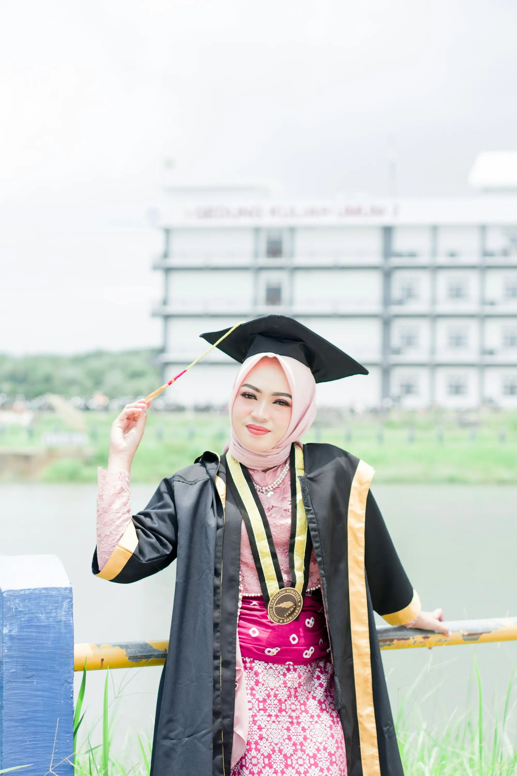 Wisudawati bertoga berpose di halaman kampus, gedung kuliah di belakang