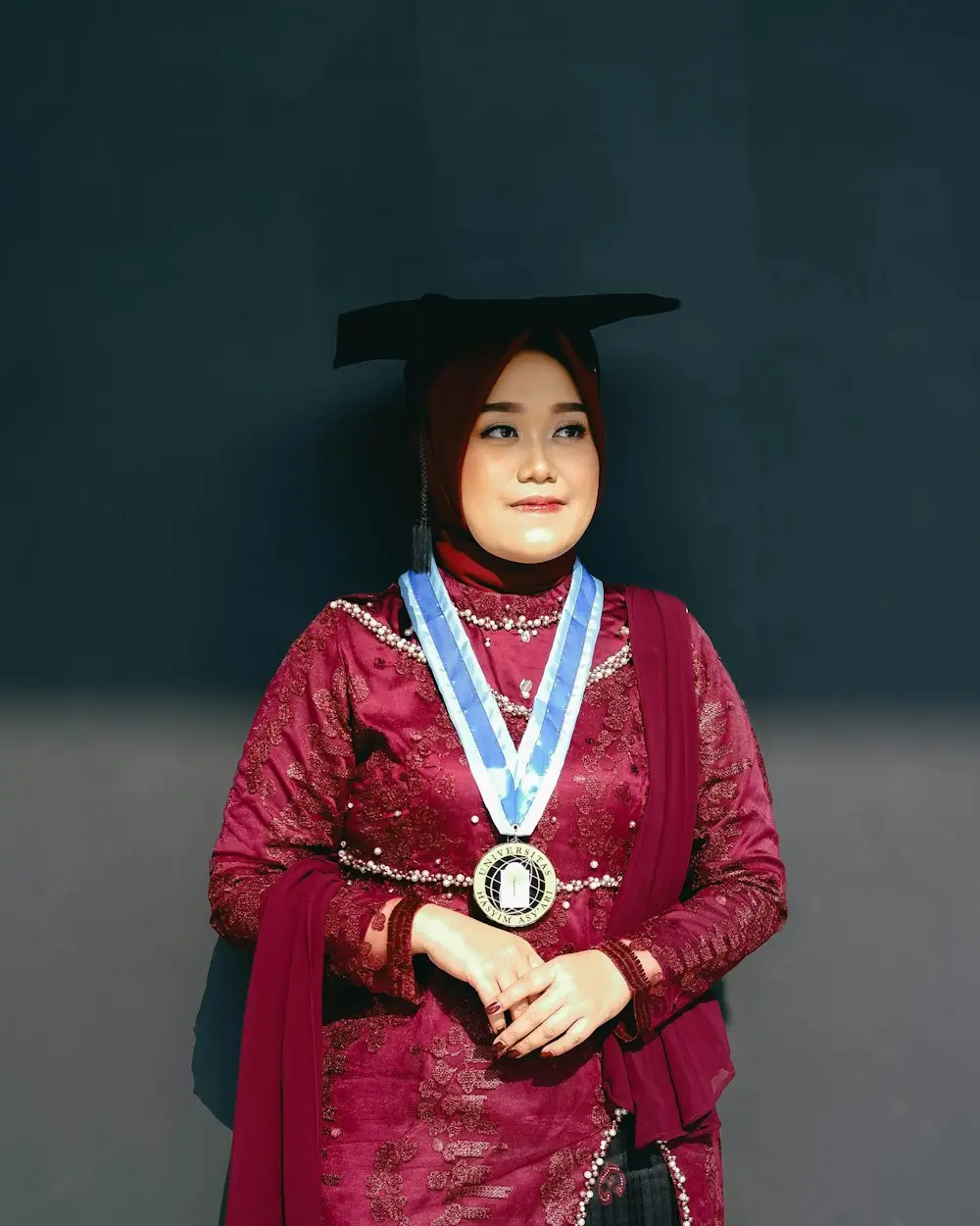 Wisudawati berkebaya merah marun dengan samir dan toga, latar gelap
